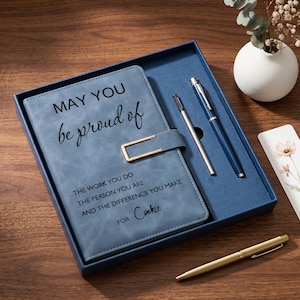 Könnte beinhalten: Ein blaues Notizbuch mit einem goldfarbenen Metallverschluss und dem Text "MAY YOU be proud of THE WORK YOU DO THE PERSON YOU ARE AND THE DIFFERENCE YOU MAKE FOR Cookie" in einer Geschenkbox mit zwei Stiften, einem blauen und einem silbernen, sowie einem Blumen-Lesezeichen.