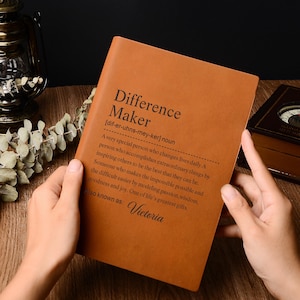 Könnte beinhalten: Ein braunes Lederjournal mit dem Text "Difference Maker" und einer Definition. Das Journal ist geöffnet und zeigt die Definition. Der Name "Victoria" ist ebenfalls auf dem Journal aufgedruckt.
