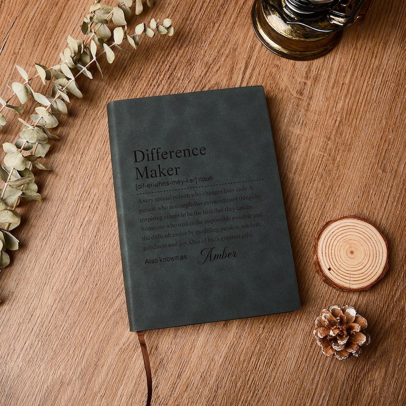 Op de afbeelding: Een donkergrijs notitieboek met de tekst "Difference Maker" en een definitie. Het notitieboek heeft een bruin lint als bladwijzer en de naam "Amber" onderaan. Het ligt op een houten oppervlak met decoratieve elementen.