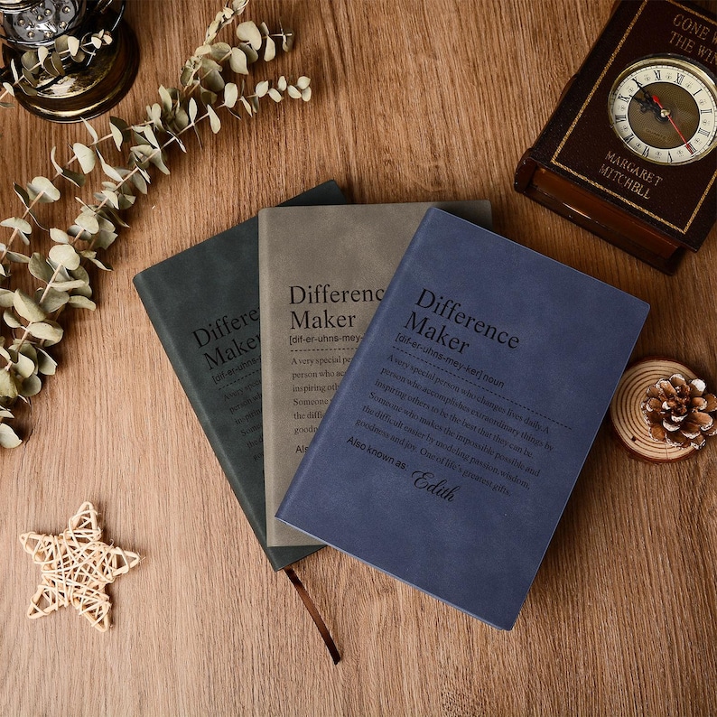 Op de afbeelding: Drie hardcover-tijdschriften in tinten blauw, grijs en groen, met de titel "Difference Maker" op de omslagen. De tijdschriften hebben een leerachtige textuur en bevatten een definitie. Een vintage klok en decoratieve items staan op de achtergrond.