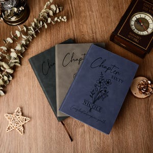 May include: Three hardback journals in shades of blue, gray, and green are displayed on a wooden surface. The blue journal is inscribed with "Chapter Sixty" and "Nicole". A clock and decorative items are in the background.