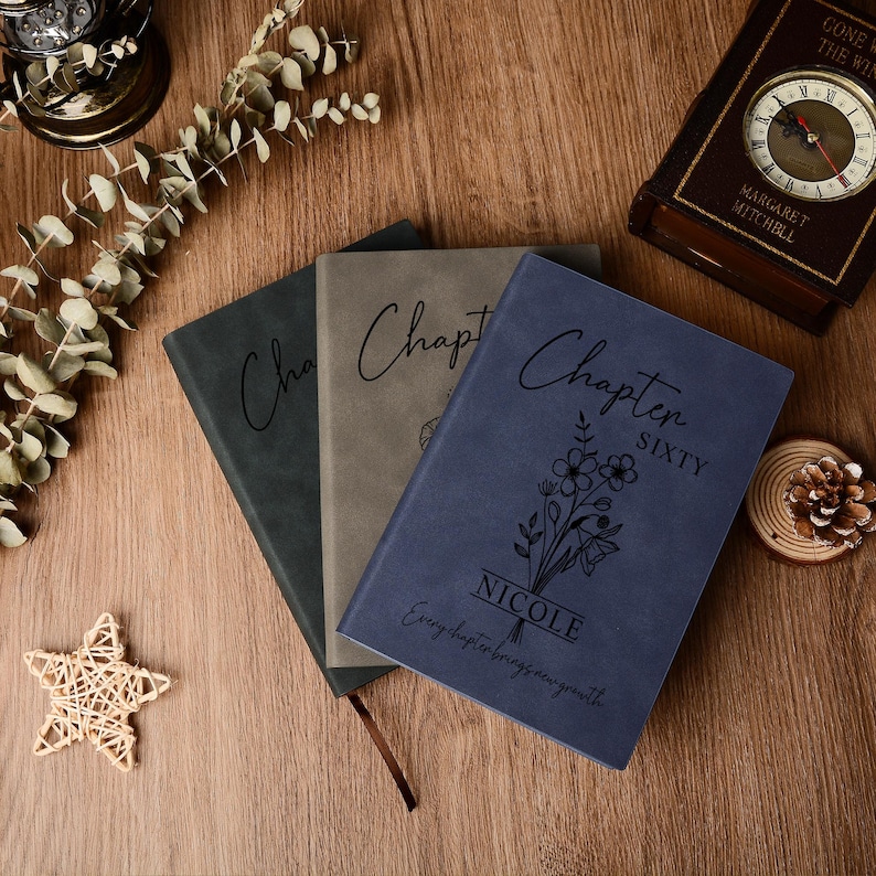May include: Three hardback journals in shades of blue, gray, and green are displayed on a wooden surface. The blue journal is inscribed with "Chapter Sixty" and "Nicole". A clock and decorative items are in the background.