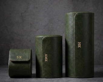 May include: Three green leather watch cases of varying sizes. The smallest case has a flap closure and the initials "SK" in gold. The middle case has the initials "AMK", and the largest has "HK". All cases are cylindrical.