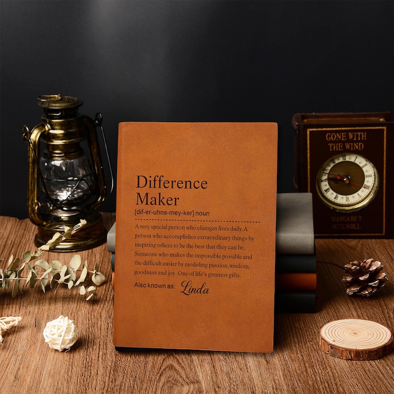 Op de afbeelding: Een bruin leren notitieboek met de tekst "Difference Maker" en een definitie. Het notitieboek ligt op een houten oppervlak met een vintage lantaarn, een klok en andere decoratieve items. Ook bekend als Linda.