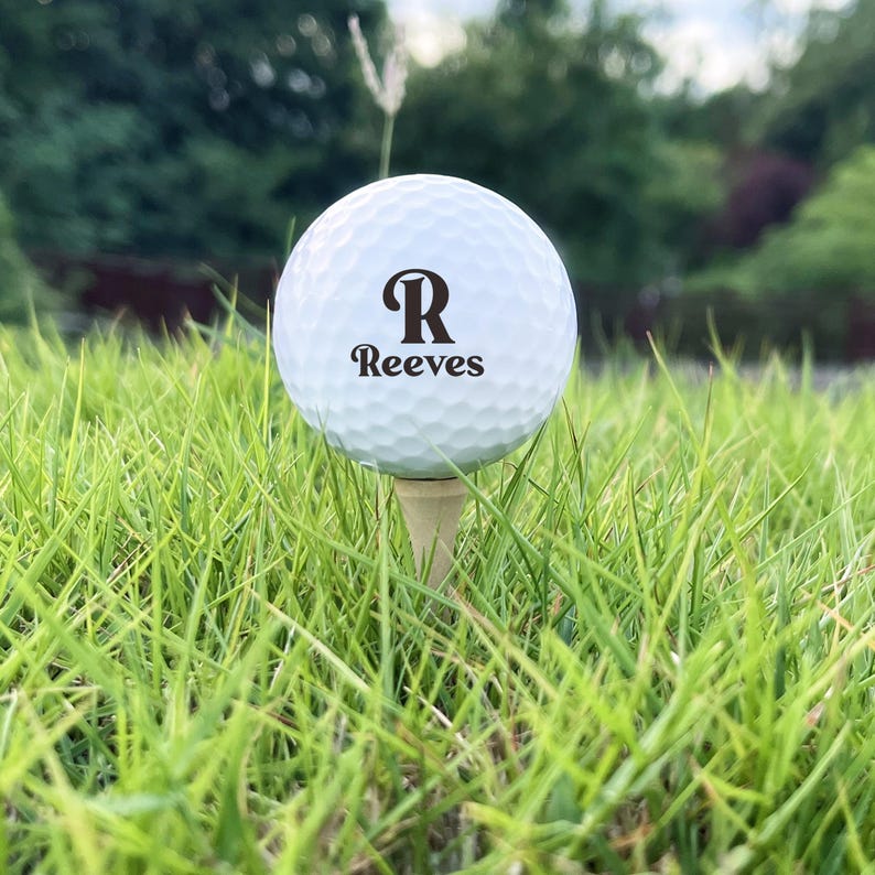 Puede incluir: Una pelota de golf blanca con el nombre "Reeves" y la letra "R" impresa, colocada sobre un tee de madera en hierba verde. El fondo muestra m&aacute;s hierba y &aacute;rboles.