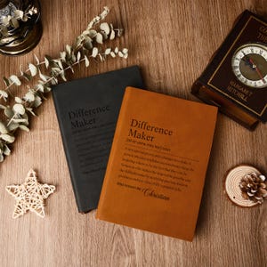 Op de afbeelding: Twee hardcover notitieboeken, &eacute;&eacute;n zwart en &eacute;&eacute;n bruin, met de tekst "Difference Maker" en een definitie gedrukt op de kaft. Een klok in vintage-stijl en decoratieve items zijn gerangschikt op een houten oppervlak.