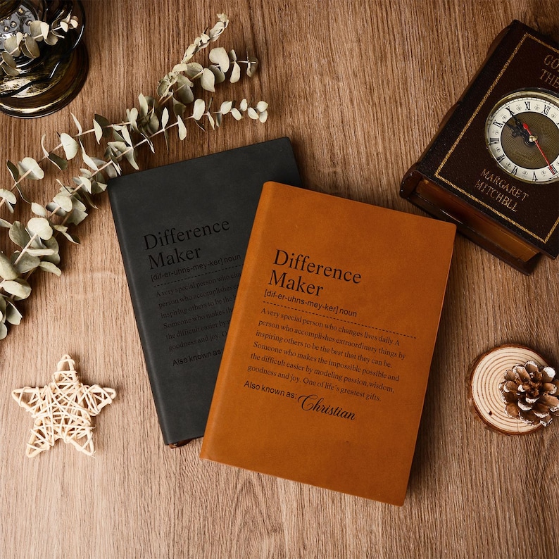 Op de afbeelding: Twee hardcover notitieboeken, &eacute;&eacute;n zwart en &eacute;&eacute;n bruin, met de tekst "Difference Maker" en een definitie gedrukt op de kaft. Een klok in vintage-stijl en decoratieve items zijn gerangschikt op een houten oppervlak.