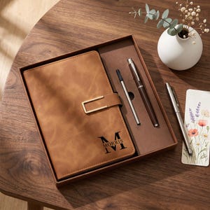 May include: A tan leather-like notebook with a gold-tone metal clasp and the name MORRIS printed in black, presented in a gift box with a dark brown pen, a silver ink refill, and a separate silver pen next to a floral illustrated bookmark.