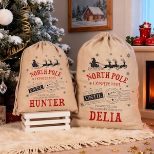 Pode incluir: Dois sacos de presente de Natal de juta bege com o texto "North Pole Express Mail" e os nomes "Hunter" e "Delia" em vermelho. Os sacos apresentam um design de Papai Noel e renas. Uma árvore de Natal e uma lareira estão no fundo.