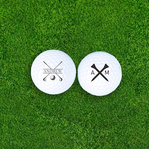 Puede incluir: Dos pelotas de golf blancas sobre hierba verde. Una bola tiene el nombre "ANDREW" con palos de golf cruzados y un gr&aacute;fico de pelota de golf. La otra bola tiene las iniciales "A" y "M" con un gr&aacute;fico de tee.