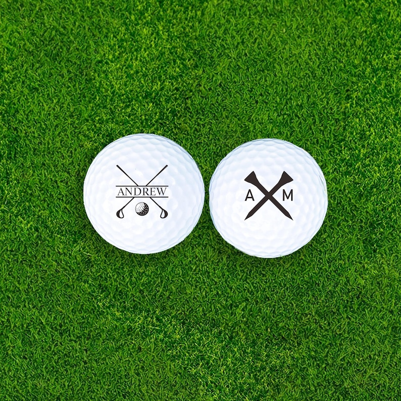 Puede incluir: Dos pelotas de golf blancas sobre hierba verde. Una bola tiene el nombre "ANDREW" con palos de golf cruzados y un gr&aacute;fico de pelota de golf. La otra bola tiene las iniciales "A" y "M" con un gr&aacute;fico de tee.