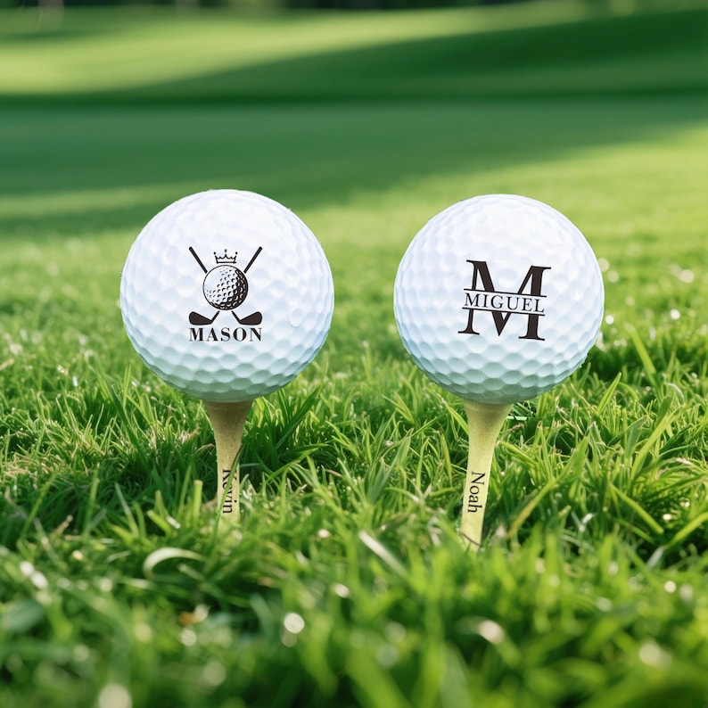 Puede incluir: Dos pelotas de golf blancas con dise&ntilde;os personalizados sobre tees en un campo de hierba. Una bola presenta un logotipo de palo y bola de golf con el nombre "Mason". La otra bola tiene la letra "M" y el nombre "Miguel". Los tees est&aacute;n grabados con nombres.