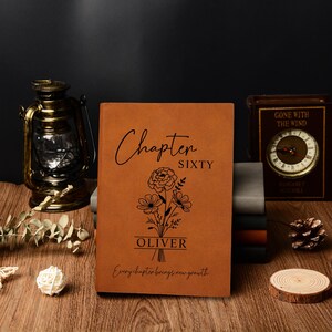 May include: A brown leather journal with the words "Chapter Sixty" and "Oliver" printed on the cover. A floral design is etched below the text. The journal is on a wooden surface with other decorative items.
