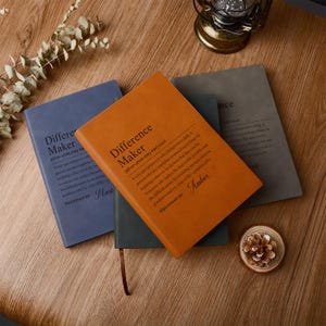 Op de afbeelding: Drie notitieboeken met harde kaft in blauw, oranje en grijs liggen op een houten oppervlak. Het oranje notitieboek op de voorgrond heeft de tekst "Difference Maker" en een definitie op de kaft.