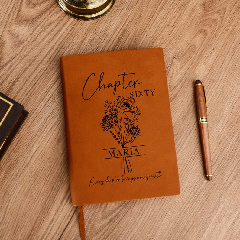 May include: A brown leather journal with the words "Chapter Sixty" and the name "Maria" printed on the cover, along with a floral design. A wooden pen rests beside the journal on a wooden surface. The text "Every chapter brings new growth" is also printed on the cover.