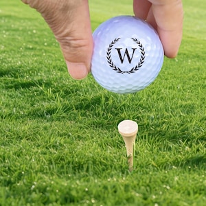 Puede incluir: Una pelota de golf blanca con una "W" negra dentro de una corona de laurel, sostenida sobre un tee en un campo de golf verde. La pelota tiene hoyuelos y est&aacute; lista para jugar.