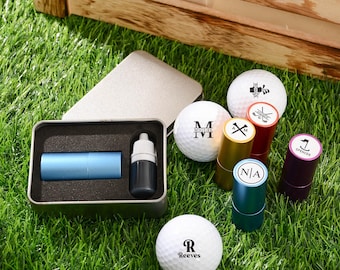 Könnte beinhalten: Eine Sammlung von Golfartikeln auf grünem Gras. Enthält personalisierte Golfbälle mit Initialen und Namen sowie verschiedenfarbiges Golfzubehör. Ein blaues Objekt und eine schwarze Flasche befinden sich in einem Metallbehälter.