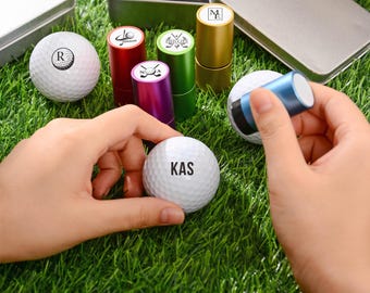 Könnte beinhalten: Weiße Golfbälle mit schwarzen Initialen und verschiedenen farbigen Golfstempeln. Die Stempel sind in Rot, Grün, Lila und Gold gehalten. Die Golfbälle und Stempel liegen auf einem grünen Kunstrasen.