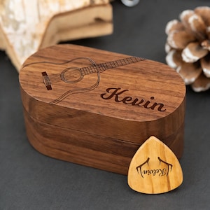 Puede incluir: Una caja de púas de guitarra de madera con un contorno de guitarra y el nombre "Kevin" grabado en la tapa. Una púa de guitarra de madera con el nombre "Kevin" y un diseño de cuerno de ciervo está al lado de la caja. La caja y la púa son de madera marrón claro.