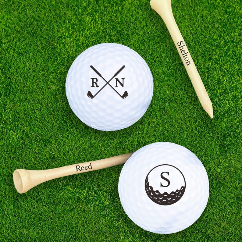 Puede incluir: Dos pelotas de golf blancas con gr&aacute;ficos negros y tees de golf de madera sobre hierba verde. Una bola tiene palos de golf cruzados con las iniciales "R" y "N". La otra bola tiene una "S" grande y un gr&aacute;fico de pelota de golf. Un tee dice "Shelton", el otro dice "Reed".