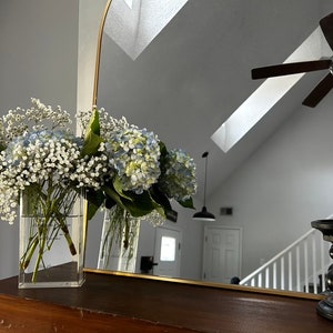 May include: A gold-framed arch mirror reflects a white ceiling with a skylight and a ceiling fan. Two clear glass vases filled with white baby's breath and blue hydrangeas sit on a wooden table in front of the mirror. Two black candle holders are on the table.