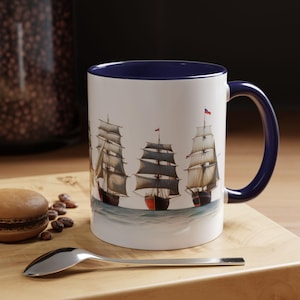 May include: White ceramic mug with a navy blue interior and handle. The mug features a watercolor illustration of several sailing ships on a blue and white background. A spoon and a pastry are in the foreground.