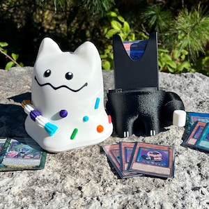 May include: A white, cat-shaped card holder with colorful sprinkles and a black, smiling mouth. A black card holder and several trading cards are also present. The items are displayed on a stone surface.