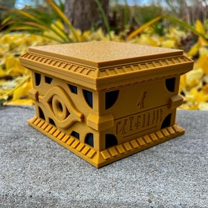 May include: A golden box with an eye symbol and hieroglyphics on the front. The box is made of plastic and has a textured surface.