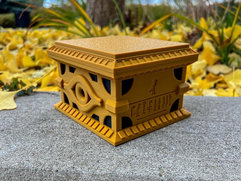 May include: A golden box with an eye symbol and hieroglyphics on the front. The box is made of plastic and has a textured surface.