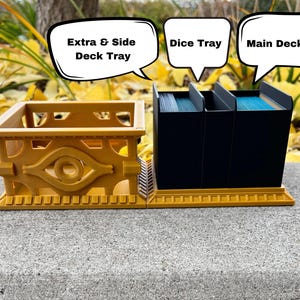 May include: A gold-colored card game organizer with three compartments for cards and a smaller compartment for dice. The compartments are labeled "Main Deck Tray", "Dice Tray", and "Extra & Side Deck Tray".