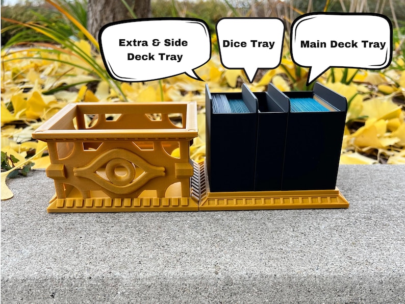 May include: A gold-colored card game organizer with three compartments for cards and a smaller compartment for dice. The compartments are labeled "Main Deck Tray", "Dice Tray", and "Extra & Side Deck Tray".