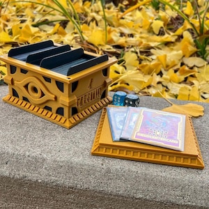 May include: A gold-colored, 3D printed deck box featuring the Eye of Horus and the text "Millennium" with a blue and black interior. The box is sitting on a gray surface with three blue dice and three trading cards with the text "Monster Card" on the bottom right corner.