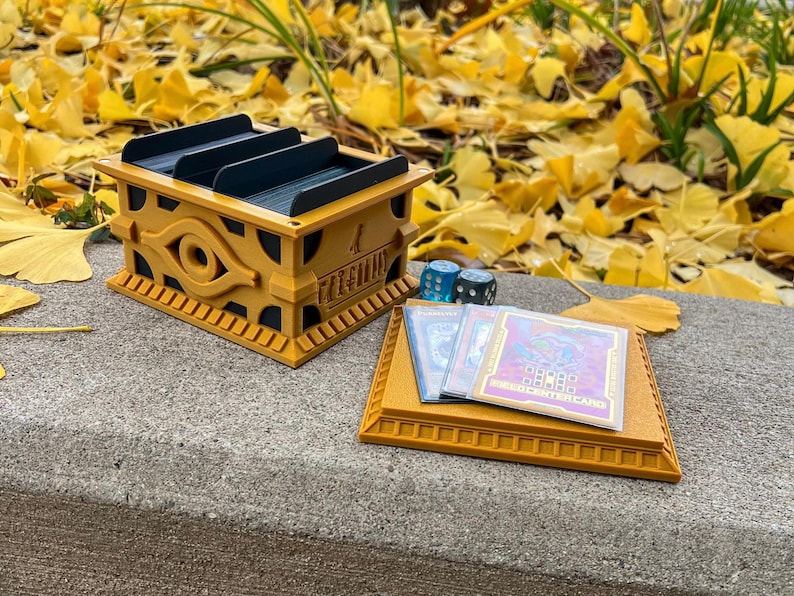 May include: A gold-colored, 3D printed deck box featuring the Eye of Horus and the text "Millennium" with a blue and black interior. The box is sitting on a gray surface with three blue dice and three trading cards with the text "Monster Card" on the bottom right corner.