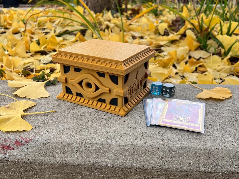 May include: A golden deck box with an eye of horus design. The box is sitting on a concrete surface with a few fallen leaves around it. There are two blue dice and a stack of trading cards next to the box.