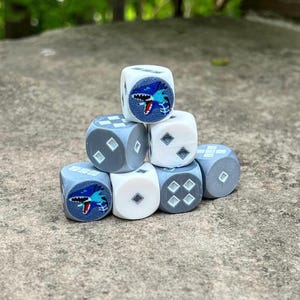 Può includere: Una collezione di sette dadi, impilati a forma di piramide. I dadi sono bianchi e grigi, con una grafica di squalo su alcune facce e motivi a diamante su altre. I dadi sono appoggiati su una superficie di pietra.