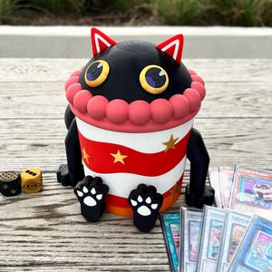 Peut inclure: Un récipient noir en forme de chat avec un motif à rayures rouges et blanches, des étoiles dorées et des pieds en forme de pattes. Le récipient a une bordure rose et est entouré de cartes à collectionner et de dés.