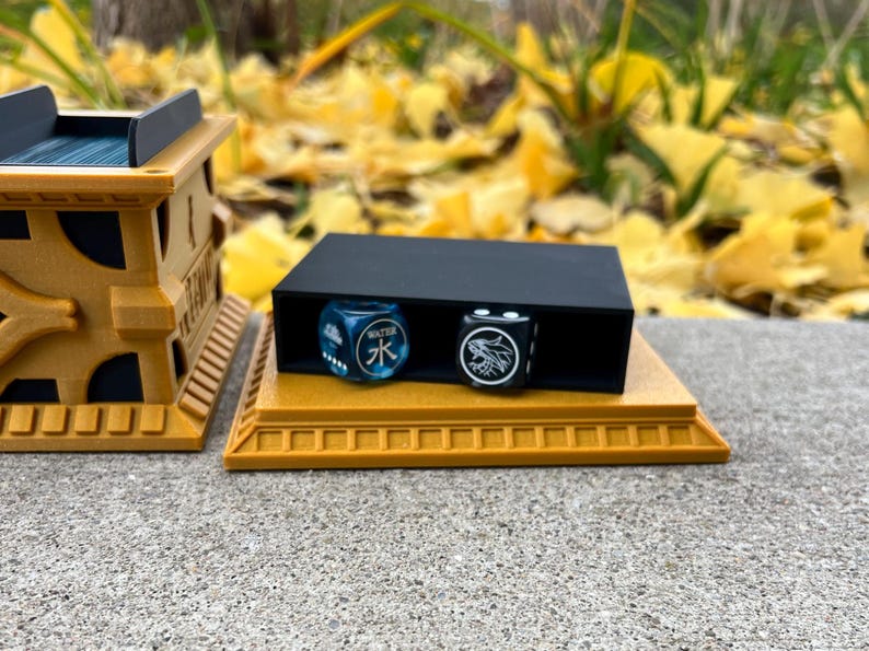 May include: A black dice tray with two dice, one blue with the Chinese character for water and the other black with a white wolf design, sits on a gold-colored base.