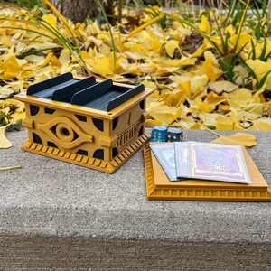 May include: A gold-colored card game deck box with a raised eye of horus design. The box is open and has three slots for cards. There are also three dice and two cards on a gold-colored tray.
