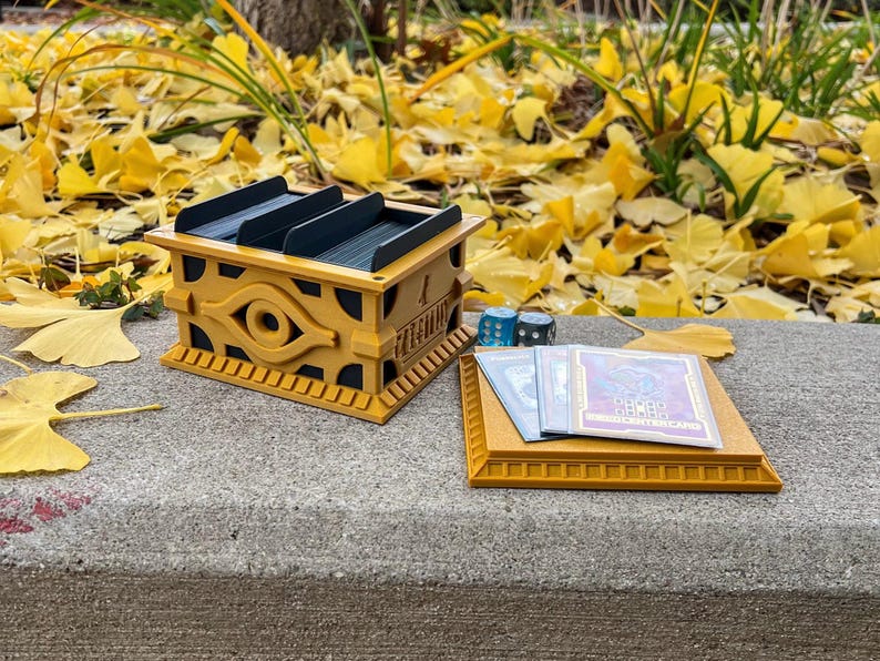 May include: A gold-colored card game deck box with a raised eye of horus design. The box is open and has three slots for cards. There are also three dice and two cards on a gold-colored tray.