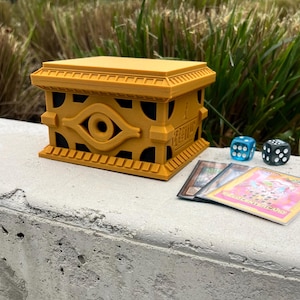 May include: A golden yellow card game deck box with a black eye of horus design. The box is sitting on a gray concrete surface with two blue dice and three playing cards.