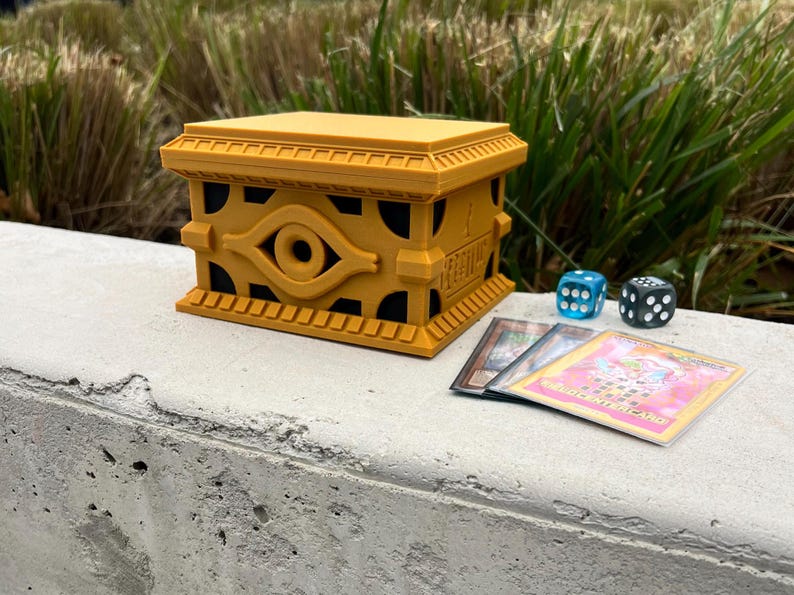May include: A golden yellow card game deck box with a black eye of horus design. The box is sitting on a gray concrete surface with two blue dice and three playing cards.