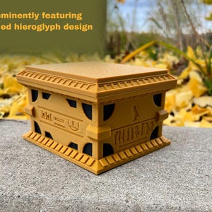 May include: A small, golden box with intricate hieroglyphic designs. The box has a raised lid and a detailed base with a small figure on the front.