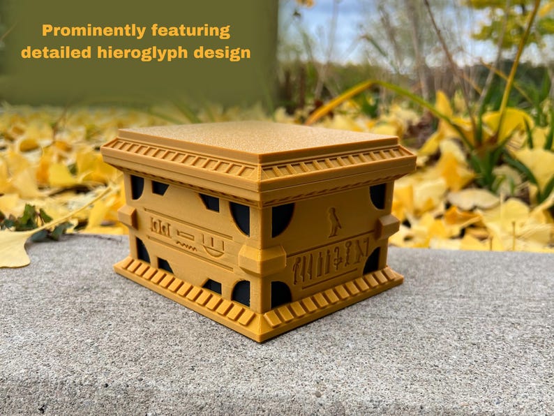 May include: A small, golden box with intricate hieroglyphic designs. The box has a raised lid and a detailed base with a small figure on the front.
