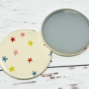May include: A round compact mirror with a white background and a pattern of colorful stars. The mirror is closed, showing the patterned side.