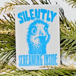 Puede incluir: Un imán rectangular blanco con las palabras "SILENTLY" y "SCREAMING INSIDE" en azul. El centro presenta una ilustración azul de la cara de un pájaro con una expresión angustiada. El imán está rodeado de follaje verde.