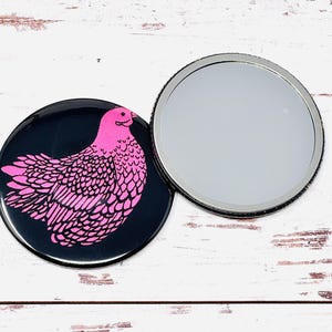 Puede incluir: Un espejo de bolsillo con fondo negro y una ilustración de pollo rosa en la parte delantera. El espejo es redondo y tiene un respaldo plateado.