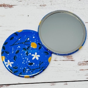 May include: A round, open compact mirror with a blue floral design. The compact features a bright blue background with white flowers, yellow lemons, and green leaves. The mirror is open, revealing the reflective surface.