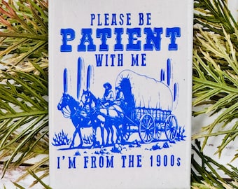 May include: A white ceramic coaster with a blue design. The design features a covered wagon pulled by two horses, with a cowboy riding in the wagon. The text on the coaster reads "Please be patient with me I'm from the 1900s".