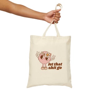 Op de afbeelding: Crèmekleurige draagtas met een cartoonillustratie. Het roze personage heeft een madeliefjeskroon en vredestekens. De tekst "let that shit go" is bruin. De tas wordt door een hand vastgehouden.