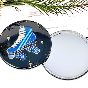 May include: A compact mirror with a black background and a blue roller skate with gold wheels. The mirror is round and has a silver rim.